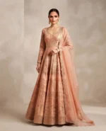 Full-length studio portrait of a model wearing a luxurious peach Anarkali suit with intricate gold embroidery and a matching sheer dupatta, set against a soft beige luxury studio background.